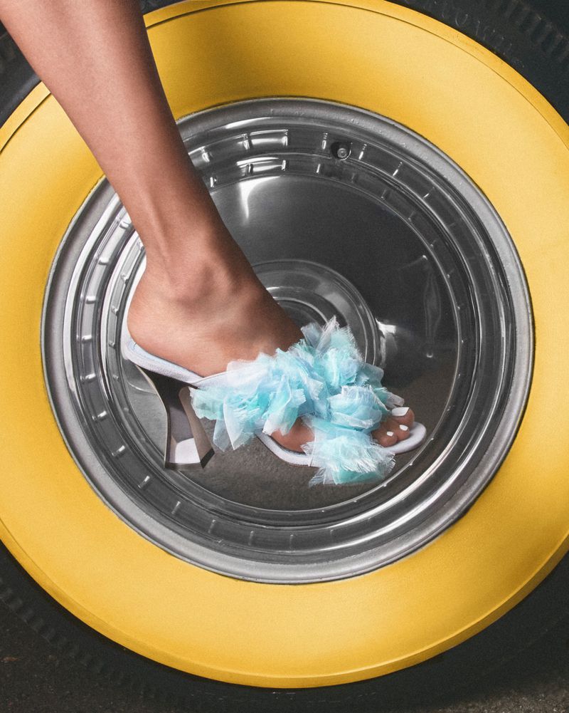 Close-up of a woman's leg and foot wearing a white heeled sandal with blue ruffled fabric on a yellow and gray tire rim.