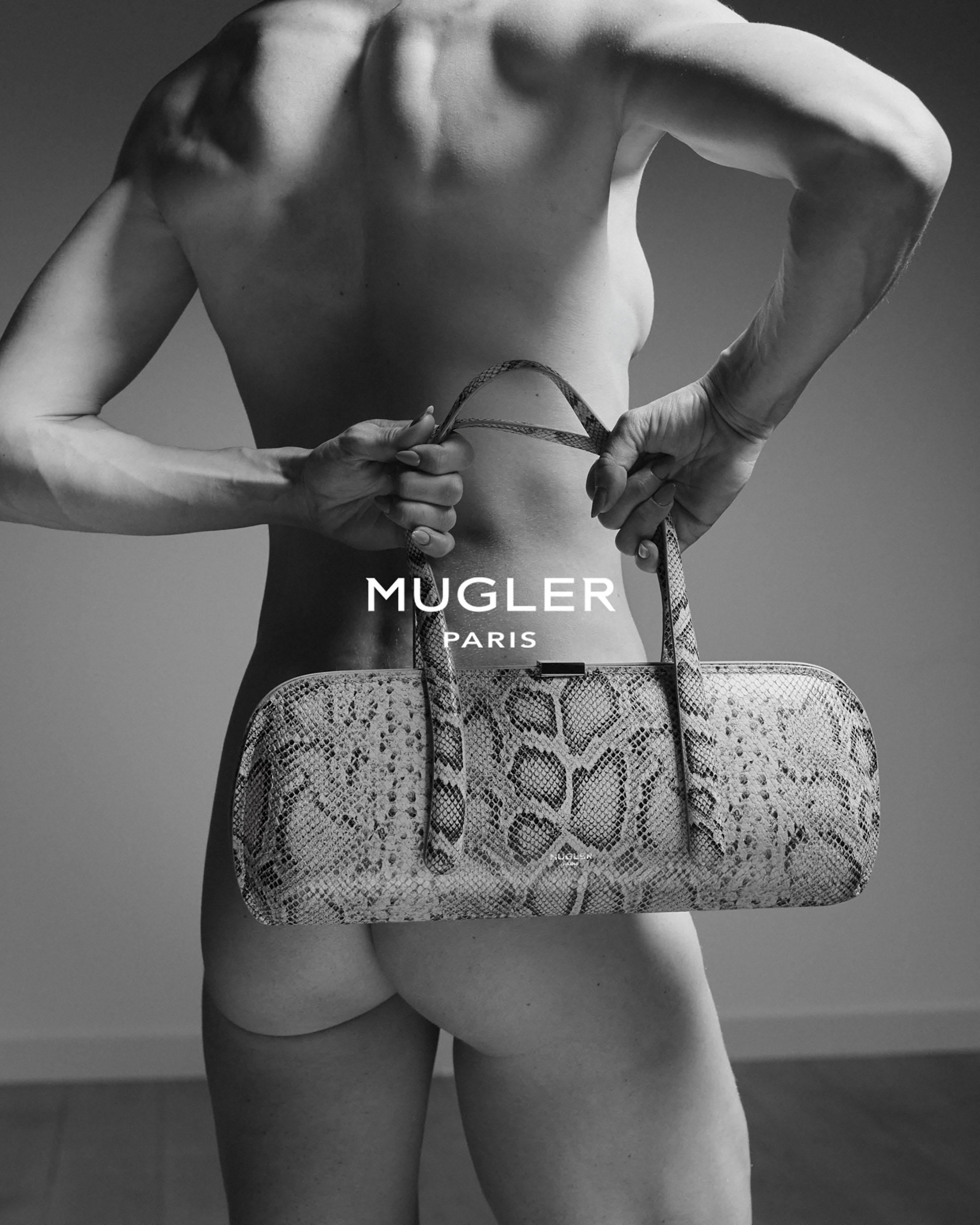 Black and white studio photograph of a muscular figure seen from behind holding an elongated snakeskin handbag with both hands.