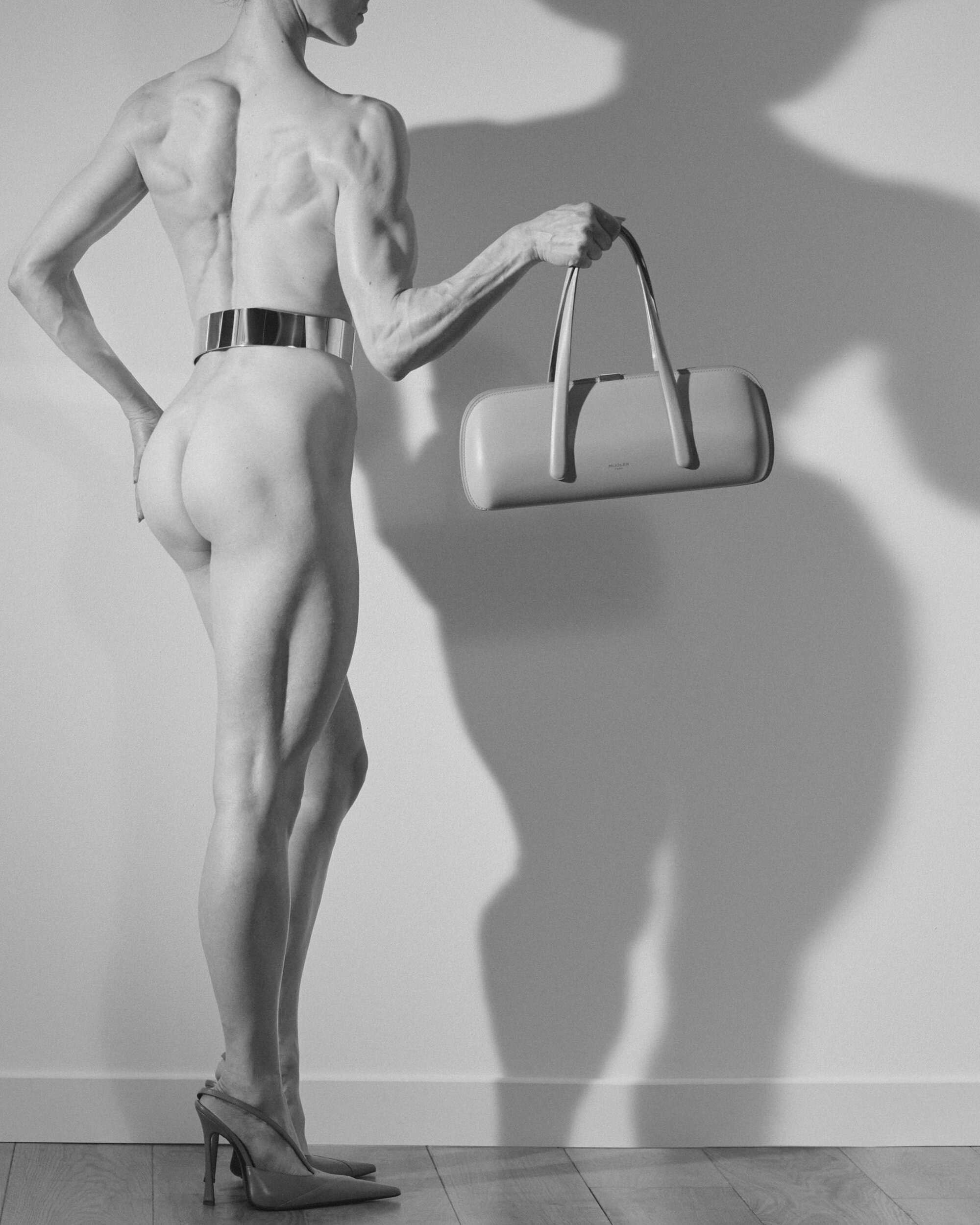 Muscular figure from behind wearing high heels and a wide metallic belt, holding a structured Mugler handbag against a studio wall.