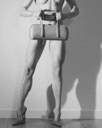 Muscular model holding a structured horizontal leather handbag while wearing pointed high-heel mules against a neutral studio wall.