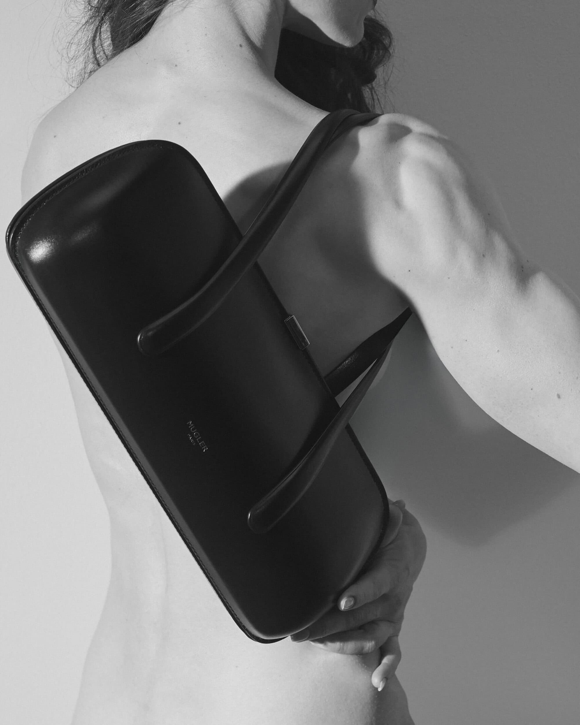 Monochrome shot of a bare-shouldered model wearing a structured black leather Mugler shoulder bag across the back against a plain backdrop.