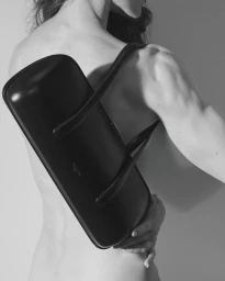 Monochrome shot of a bare-shouldered model wearing a structured black leather Mugler shoulder bag across the back against a plain backdrop.