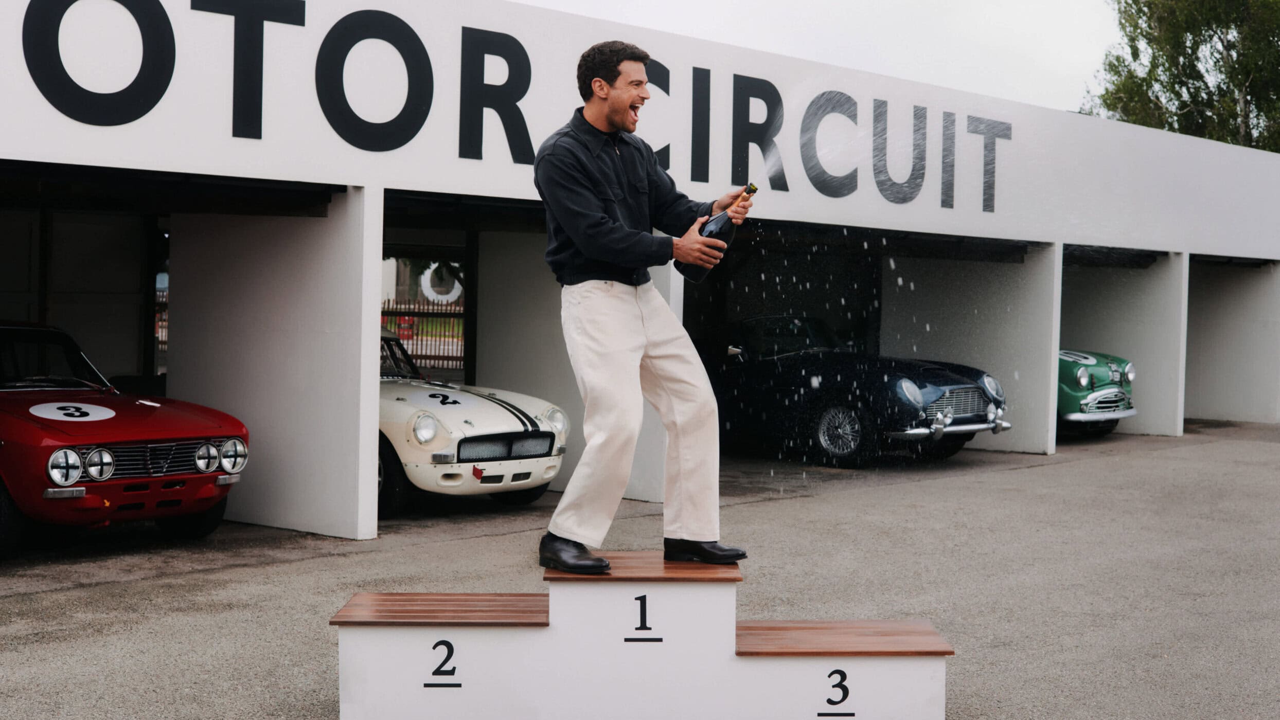 A man stands atop a first place podium spraying champagne in front of vintage race cars at a motor circuit.