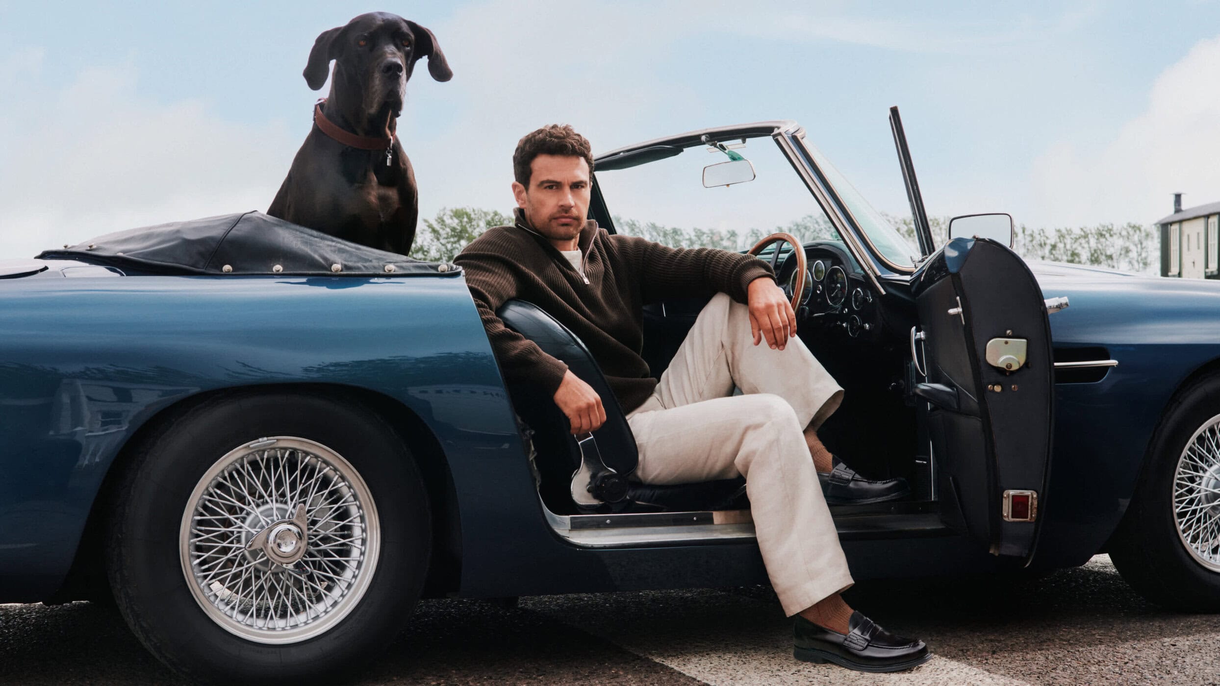 Theo James sitting in a vintage blue convertible car with a Great Dane dog in the back for Church's Fall campaign.