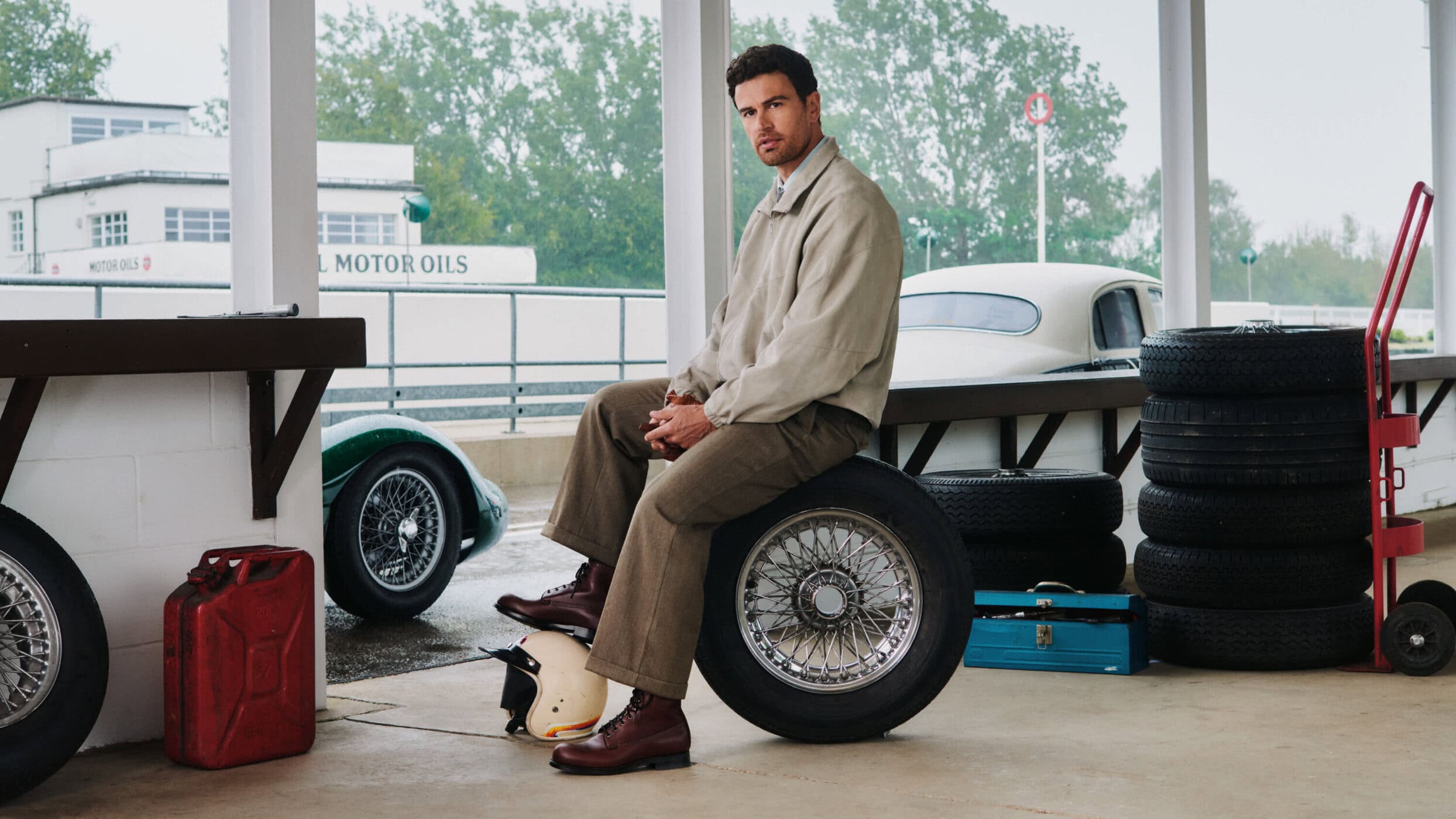 Man seated on a spare wire-wheel tire wearing a beige jacket and brown trousers in a classic automotive garage setting