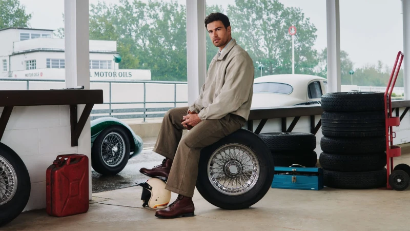 Man seated on a spare wire-wheel tire wearing a beige jacket and brown trousers in a classic automotive garage setting
