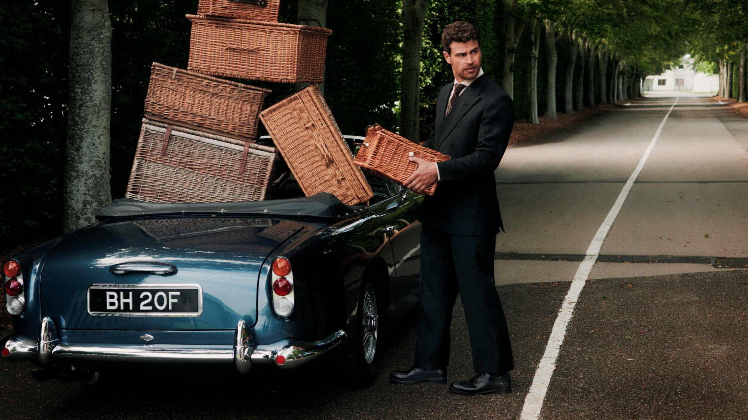 Man in a black suit holding a wicker basket beside a dark blue vintage convertible loaded with wicker baskets on a tree-lined road.
