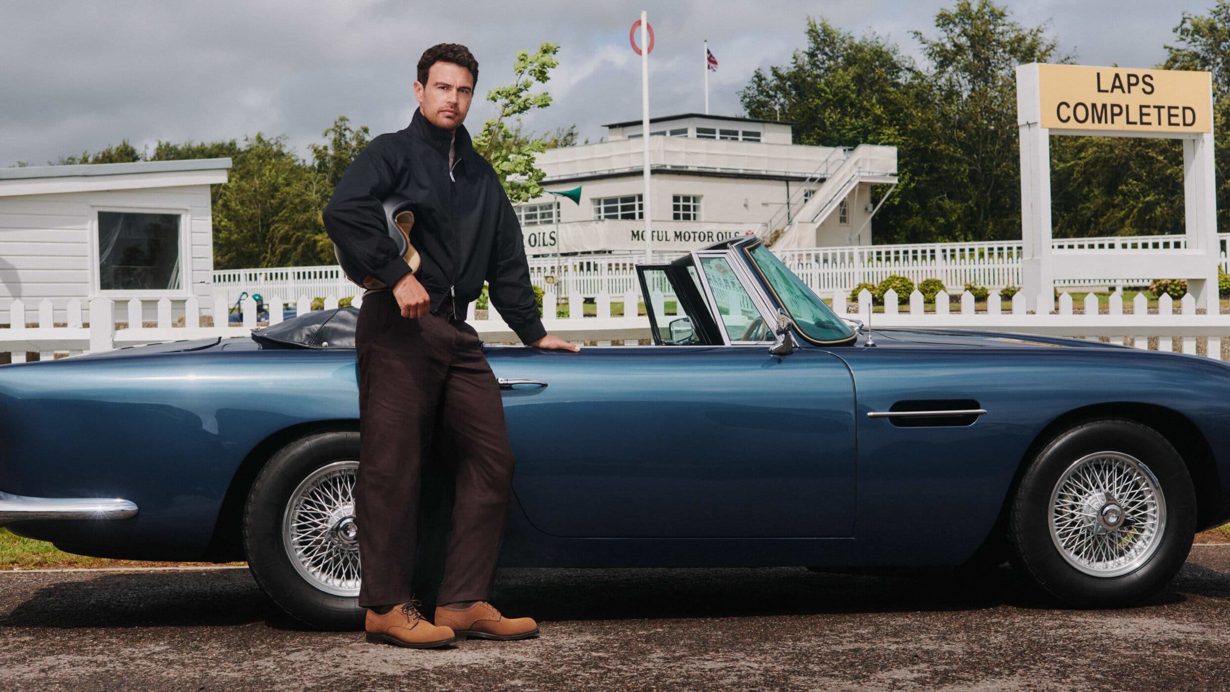 Man in a black jacket and brown trousers holding a helmet while standing beside a classic blue convertible sports car at a racetrack.