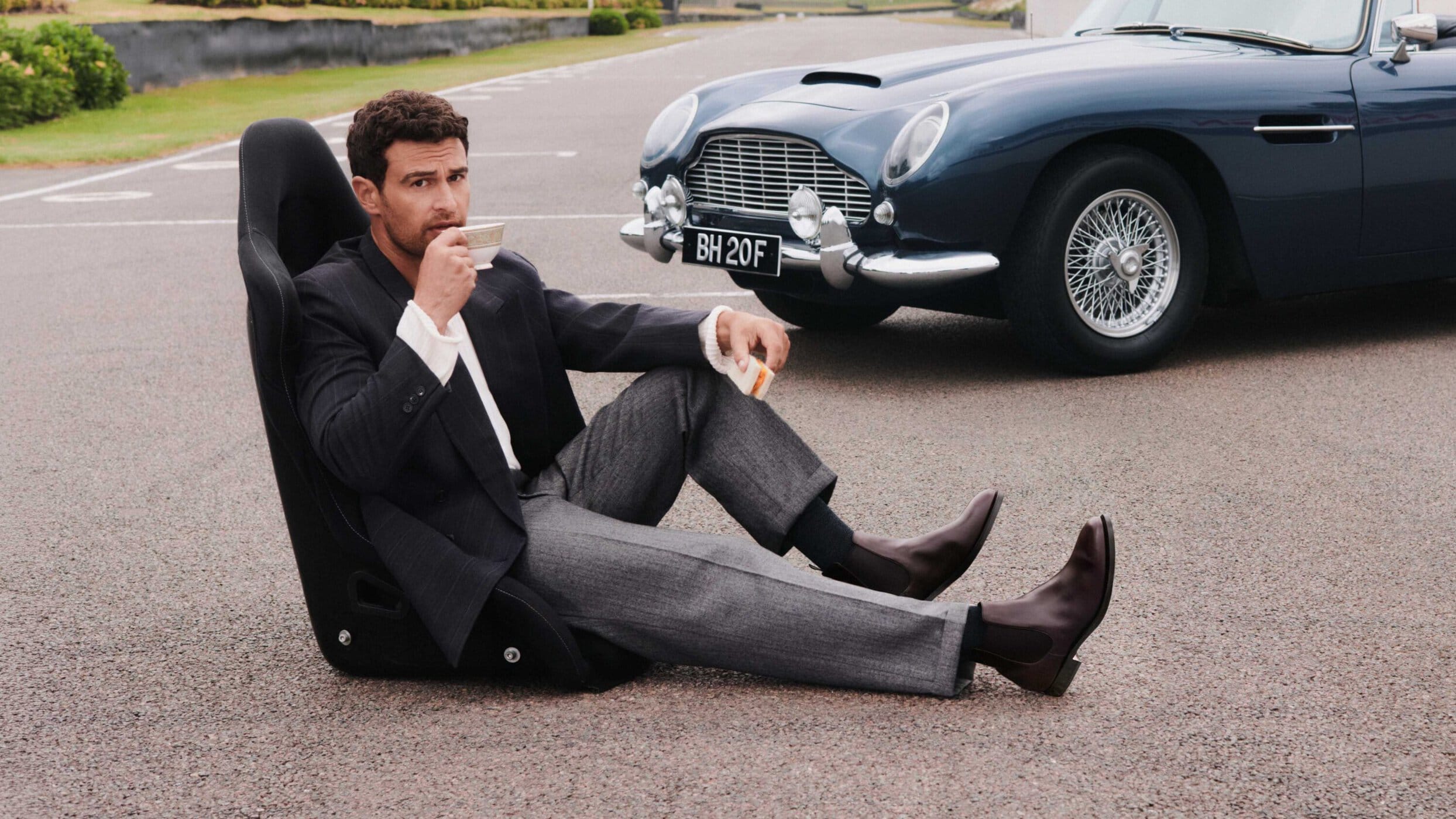 Man sitting in a detached car racing seat on an asphalt road drinking tea beside a vintage blue sports car wearing Chelsea boots.