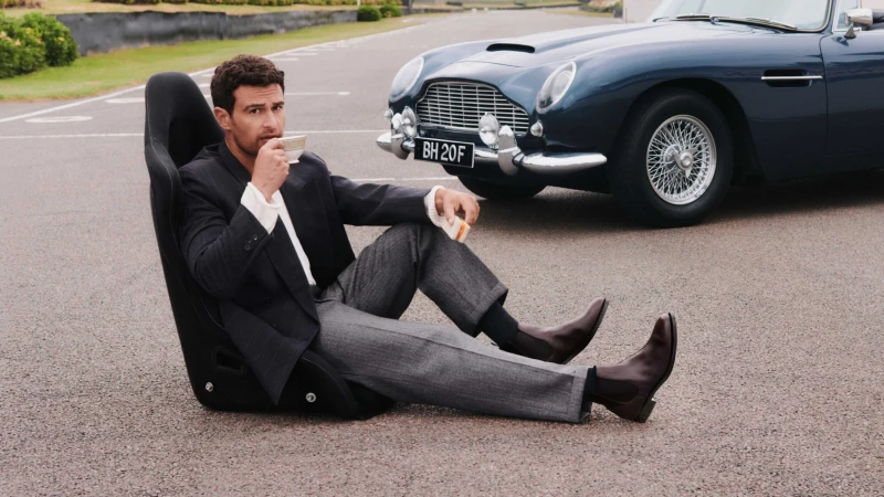 Man sitting in a detached car racing seat on an asphalt road drinking tea beside a vintage blue sports car wearing Chelsea boots.