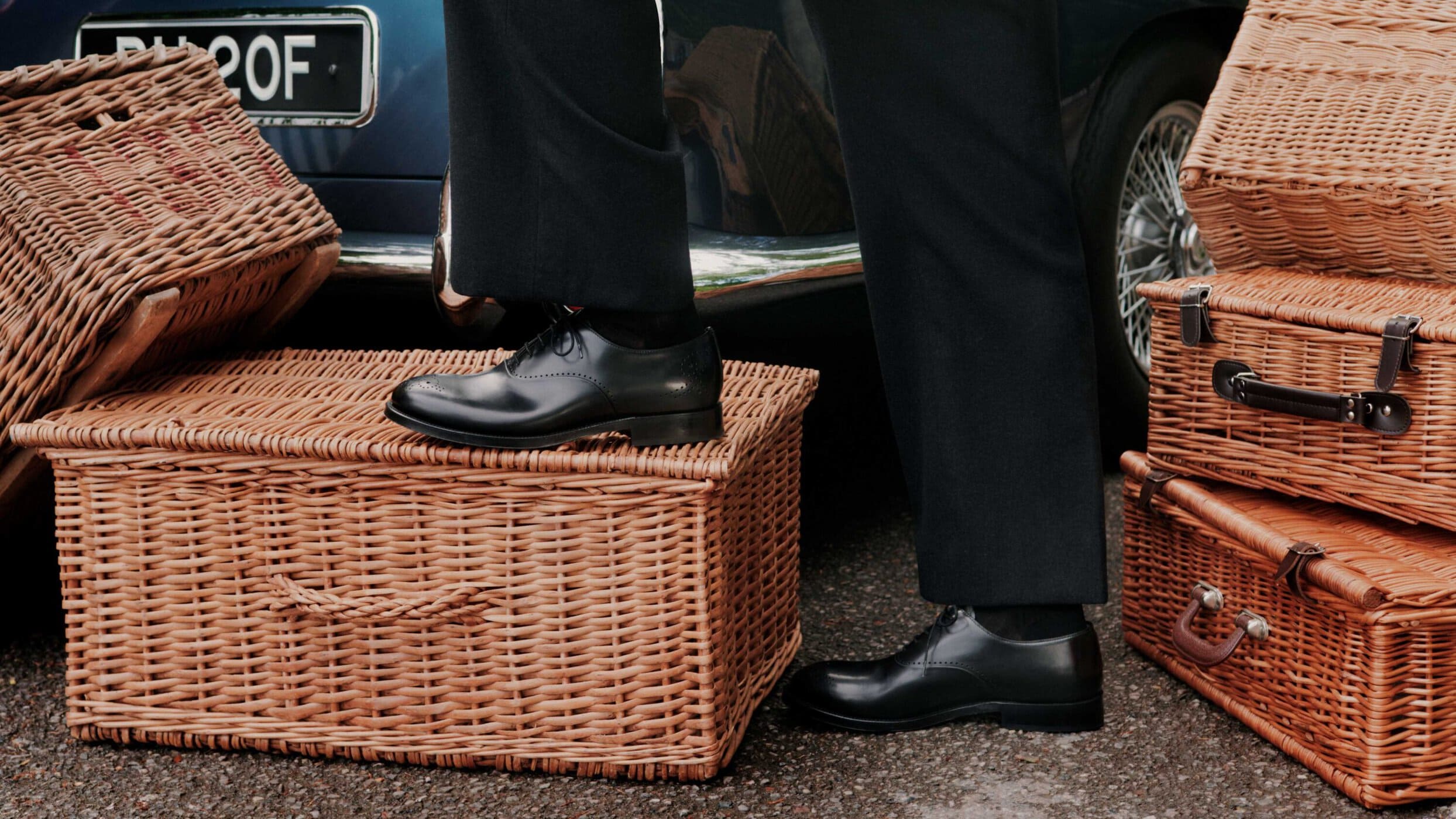 Close-up of polished black leather dress shoes resting on vintage wicker luggage beside a classic car.