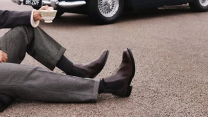 Person wearing brown leather Chelsea boots and grey wool cuffed trousers seated on asphalt holding a teacup near a vintage car.