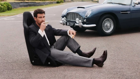 Man sitting in a detached car racing seat on an asphalt road drinking tea beside a vintage blue sports car wearing Chelsea boots.
