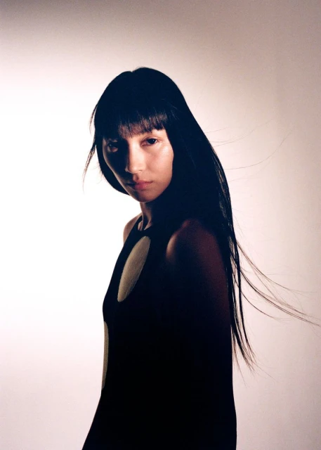 Portrait of a young Asian woman with long dark hair wearing a black sleeveless dress with circular cutout, gazing at the camera indoors.