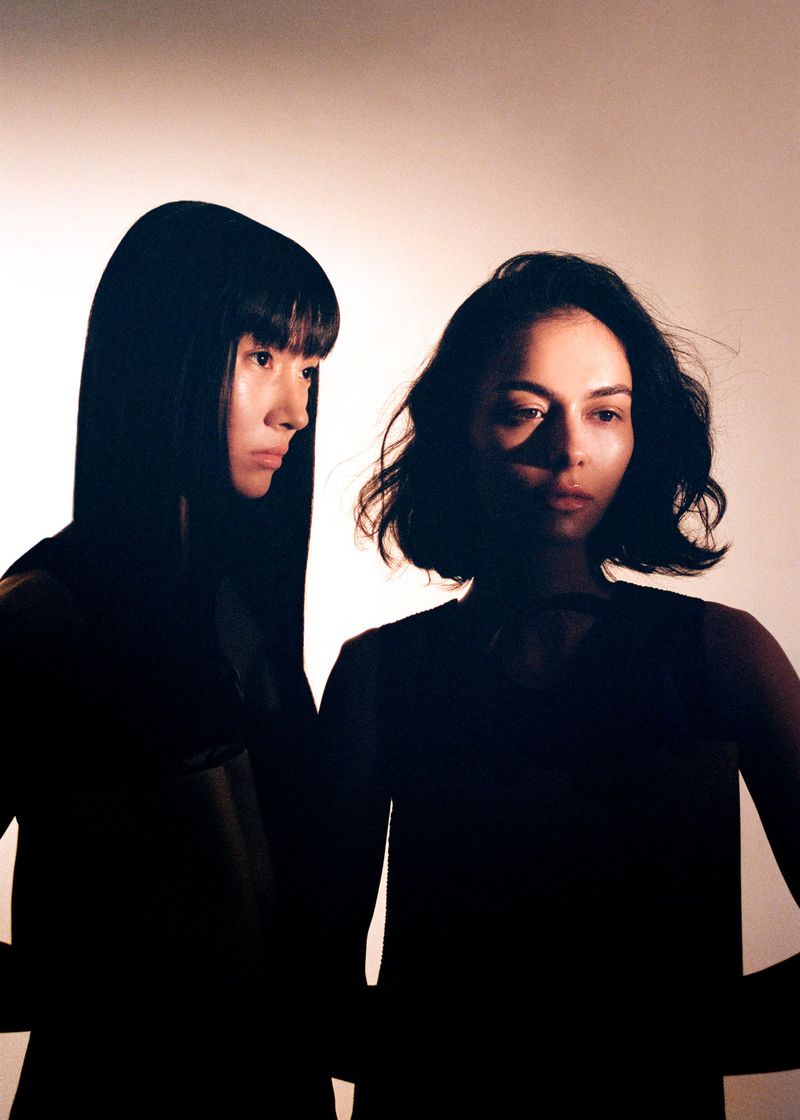 Portrait of two women in dark clothing with short and long dark hair, set against a pale backdrop with focused light, emphasizing silhouettes.