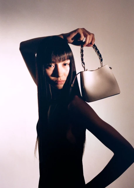 Portrait of a young woman with dark hair holding a white leather bucket bag with a chain handle, against a neutral background.