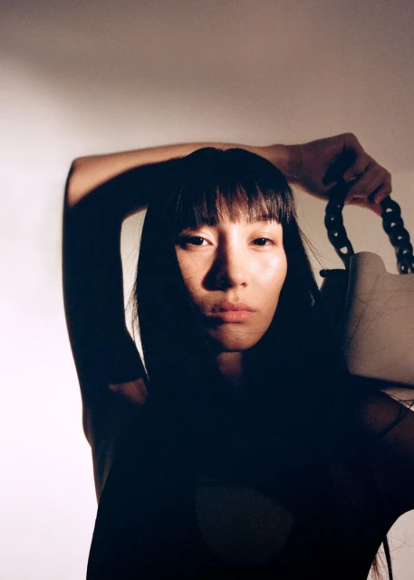 Portrait of a woman with dark hair and bangs, holding a white purse with a chunky black handle, in a studio setting with bright lighting.