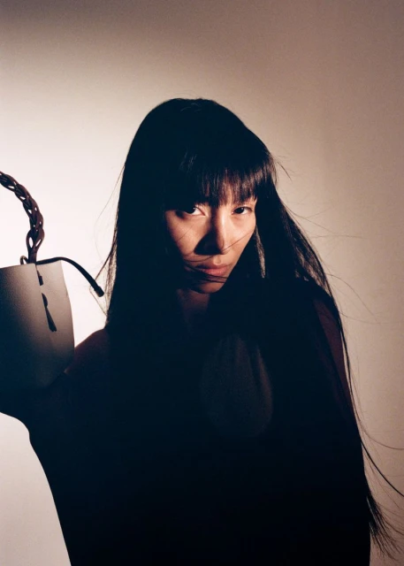Portrait of an East Asian adult woman with long, straight black hair holding a textured white bucket bag in a studio setting.
