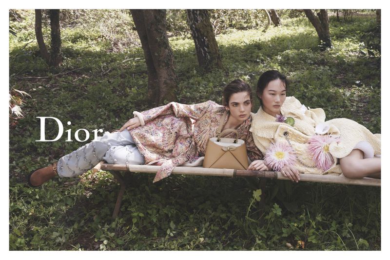 Two models reclining on a wooden cot in a wooded setting wearing patterned coats with a tan handbag between them.