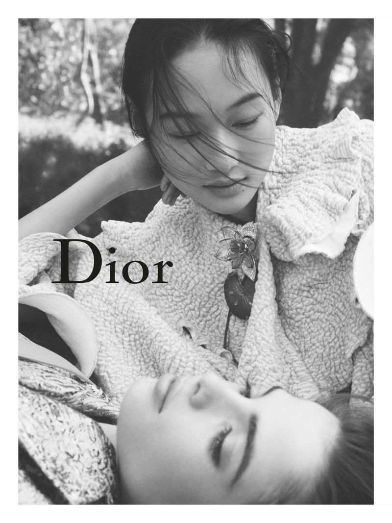 Monochrome close-up of models Zhao Ziqi and Laura Kaiser resting together wearing textured Dior garments with floral brooch.