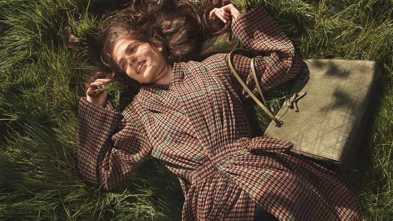 Model lying in tall green grass wearing a belted brown plaid coat next to an olive green leather tote bag.