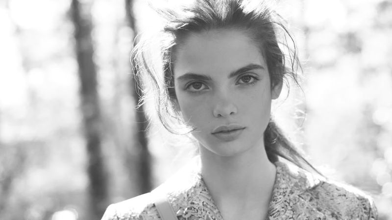 Close-up monochrome portrait of a female model outdoors with sunlight filtering through trees in the background.