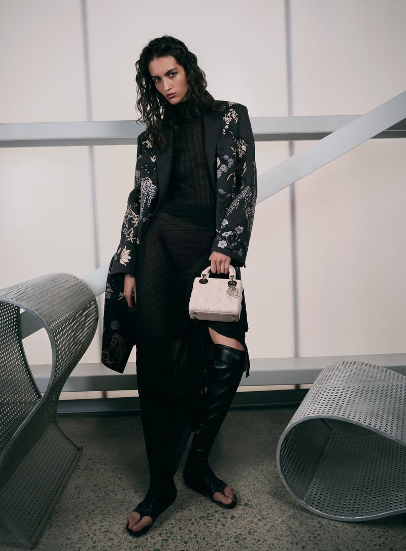 Fashion shot of a woman in a patterned coat holding a light-pink purse, posing against a white and grey background with metal details.