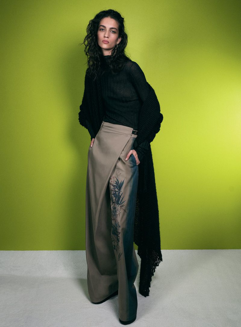 A fashion model in a black top and beige skirt poses against a light green background in a studio, showcasing contemporary style.