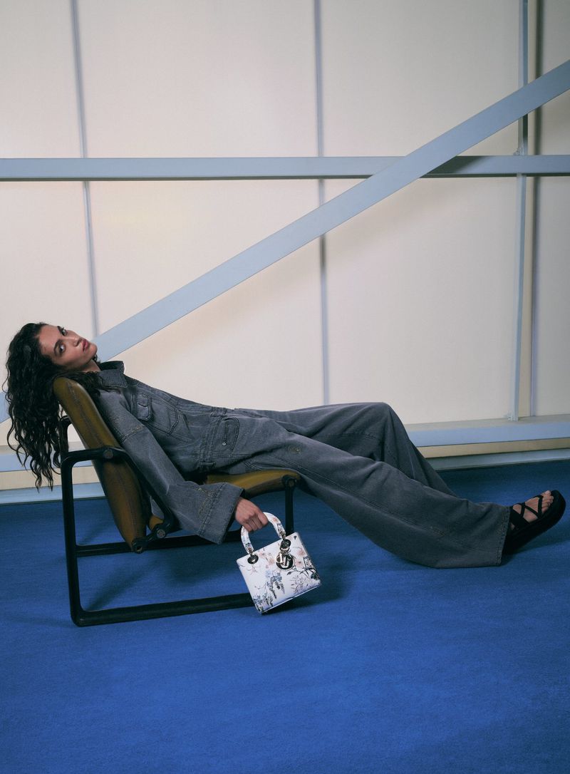 An adult female model is reclining on a chair, holding a purse, and wearing a denim outfit in a modern-style indoor environment.