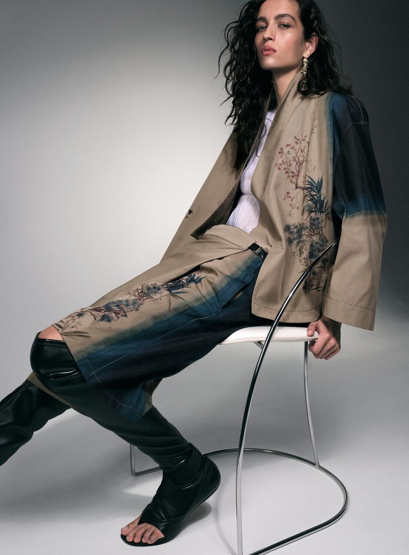 A woman with curly hair sits on a white stool in a studio wearing a jacket with painted details and high black leather sandal boots.