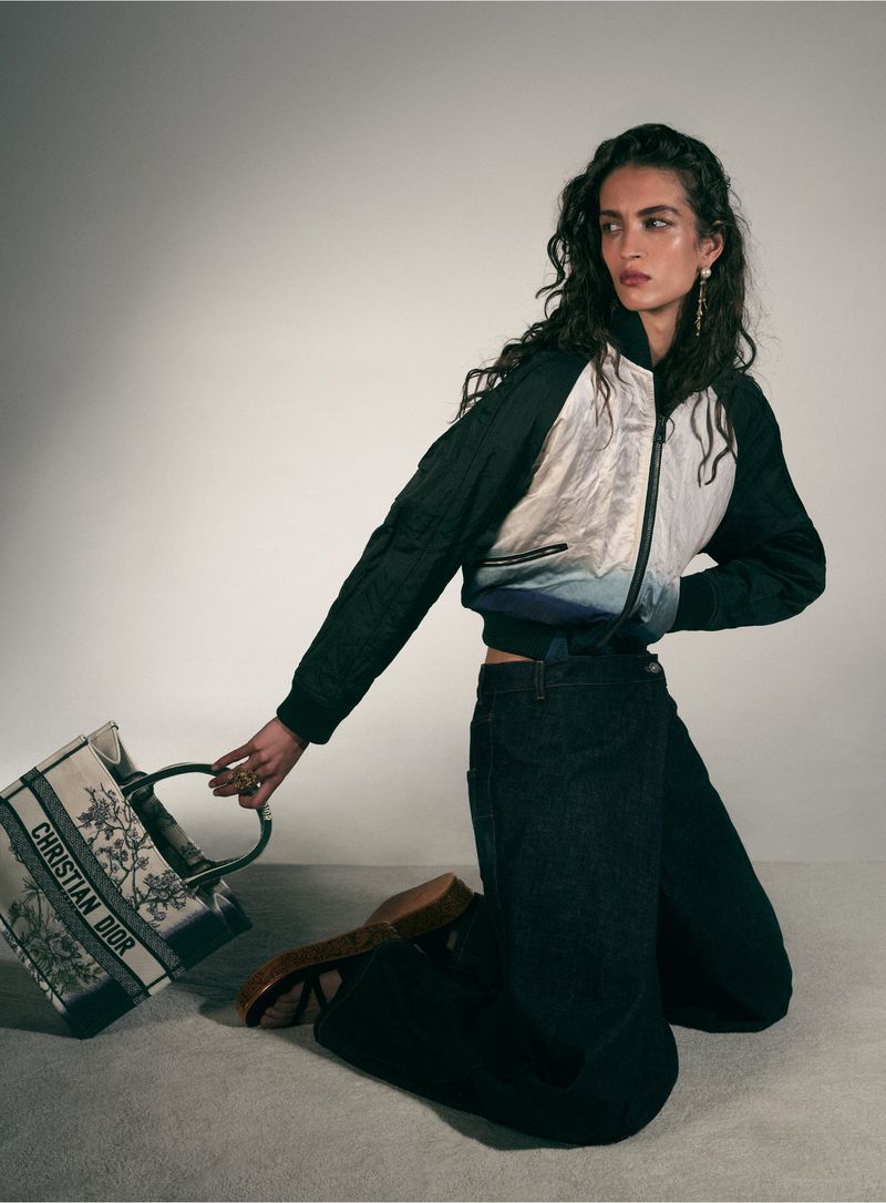 A woman kneels indoors wearing a jacket, dark jeans, and heels, holding a 'Christian Dior' tote bag against a neutral gradient backdrop.