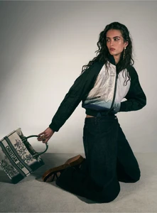 Model kneeling on a grey surface wearing a dark bomber jacket and holding a patterned tote bag against a neutral background.