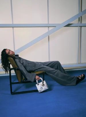 Model Lily Vander Meeden reclines in a chair holding a white printed handbag, wearing a grey denim set against a blue floor.