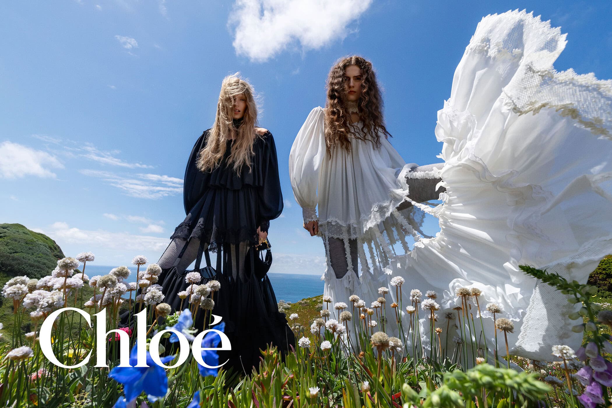 Two fashion models wearing flowing black and white ruffled gowns stand among wildflowers on a coastal hill for Chloé.