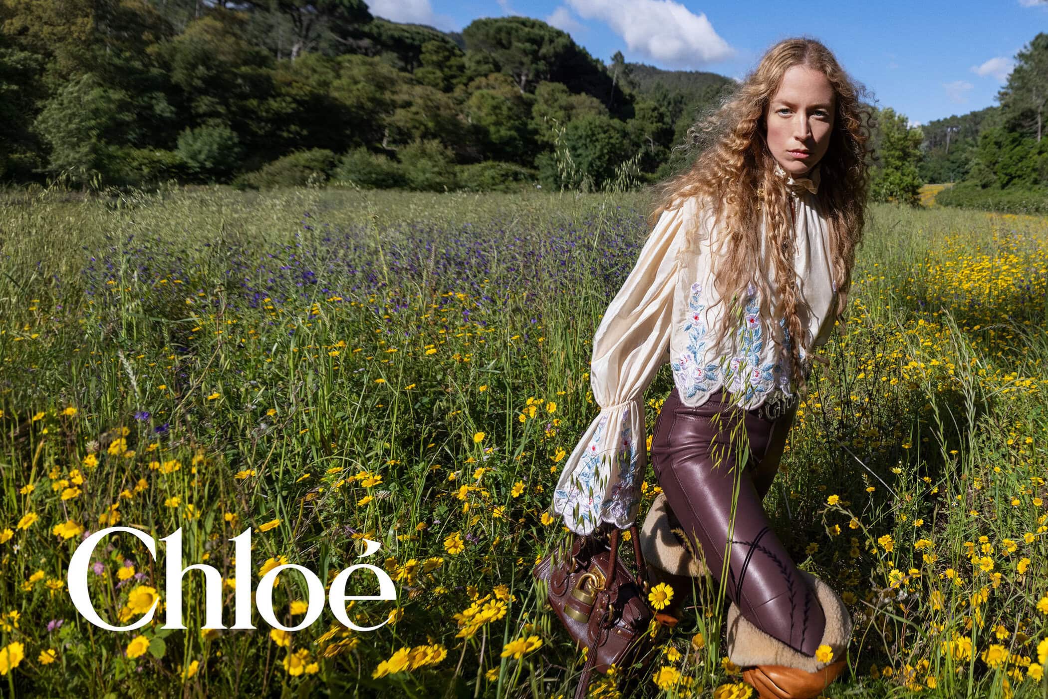 Raquel Zimmermann poses in a sunlit wildflower field wearing an embroidered cream blouse and leather pants with a brown Chloé handbag.