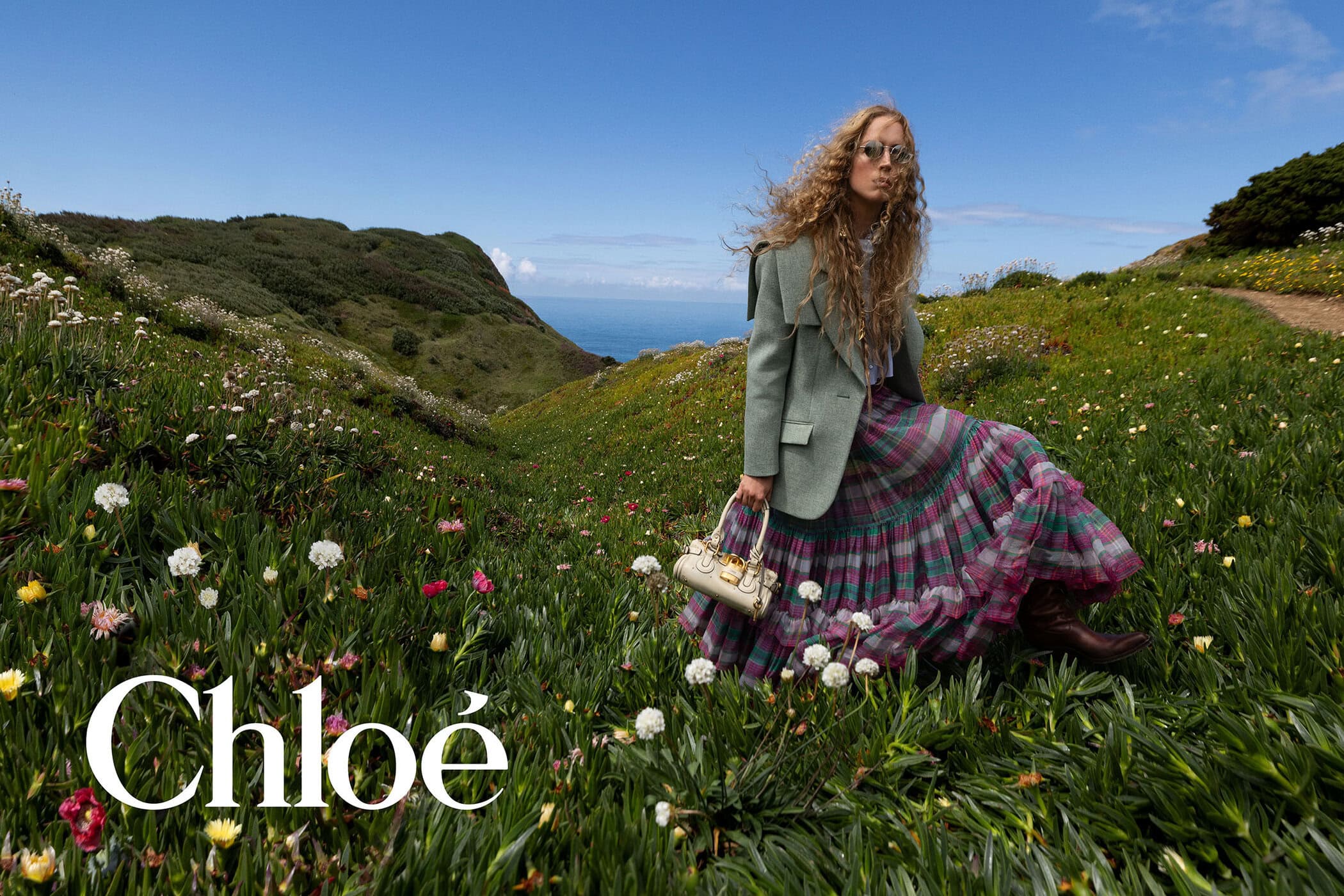 Raquel Zimmermann stands on a flowering coastal hillside wearing a sage green jacket, plaid tiered skirt, and holding a pale handbag.
