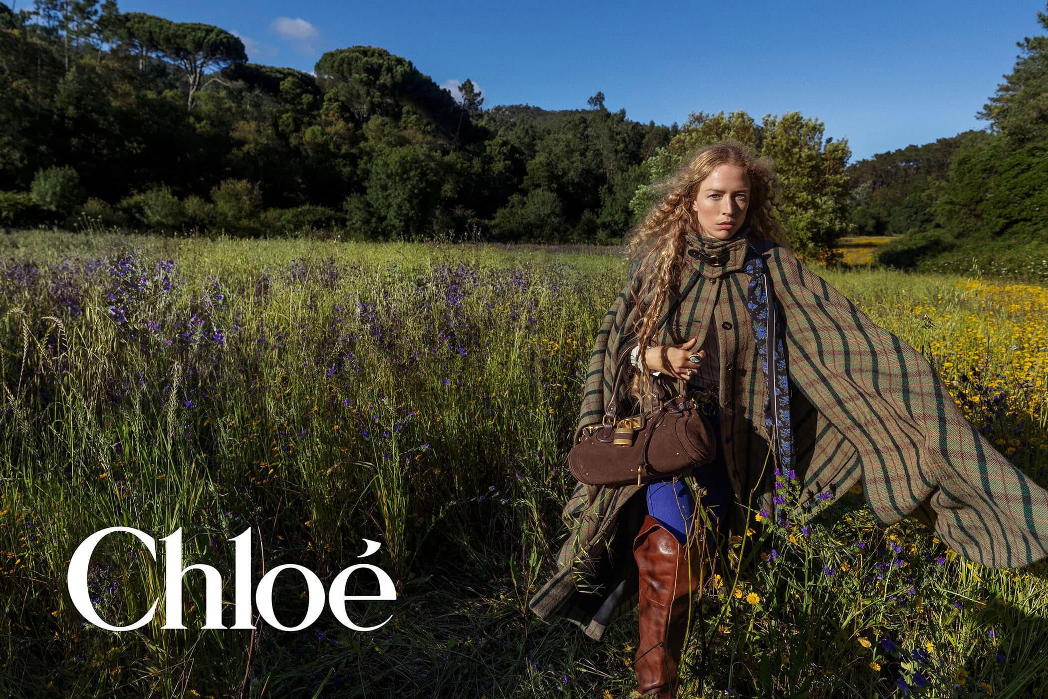 Raquel Zimmermann poses in a plaid cape coat, brown boots, and leather handbag in a grassy field for Chloé.