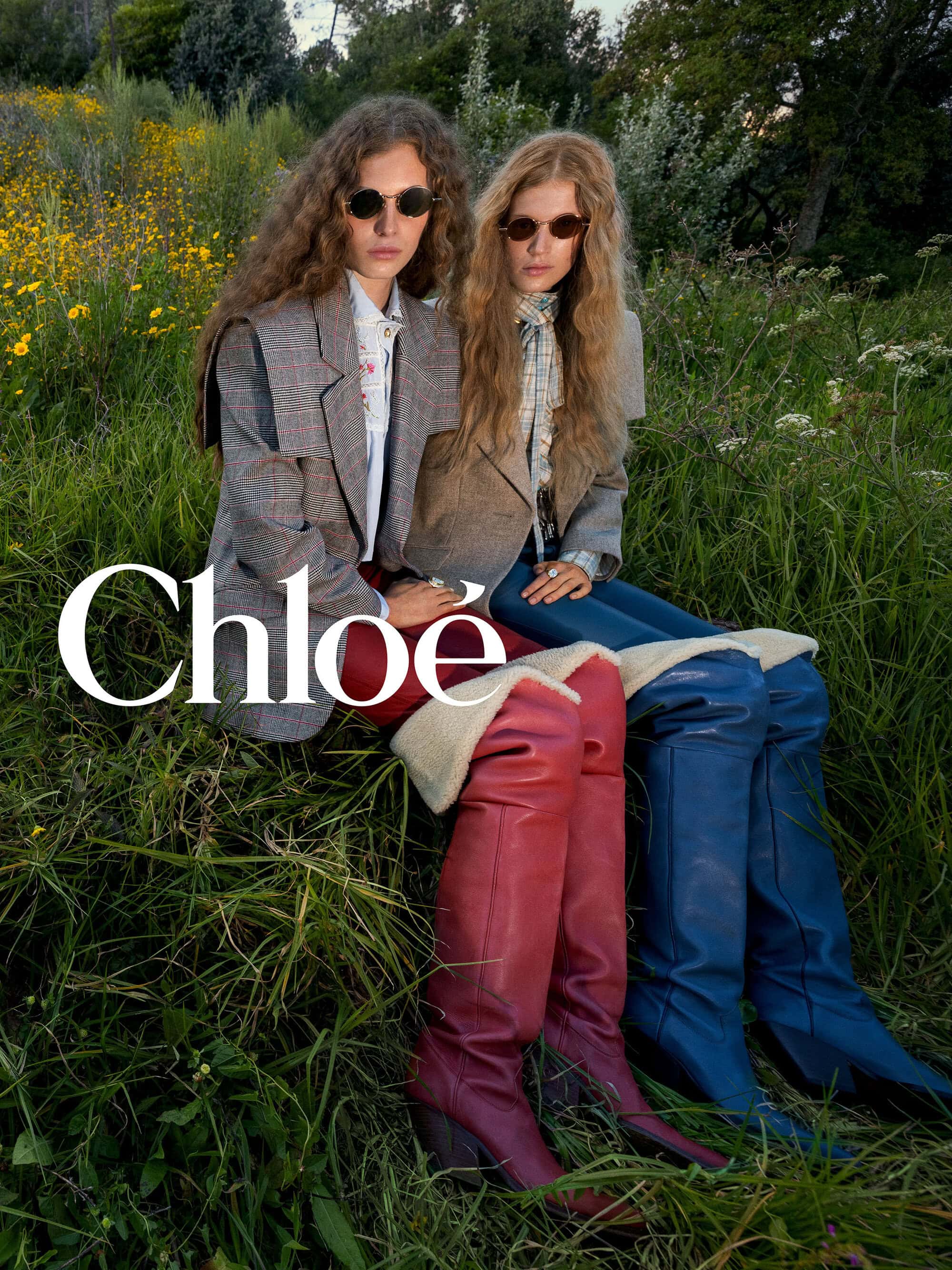 Two models sitting in a grassy wildflower meadow wearing tailored blazers, dark sunglasses, and shearling-lined leather boots for Chloé.