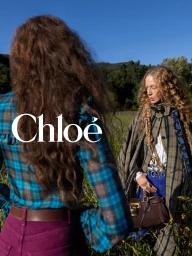 Two women with long curly hair in plaid garments and carrying a leather handbag standing in an outdoor grassy field under direct sunlight.