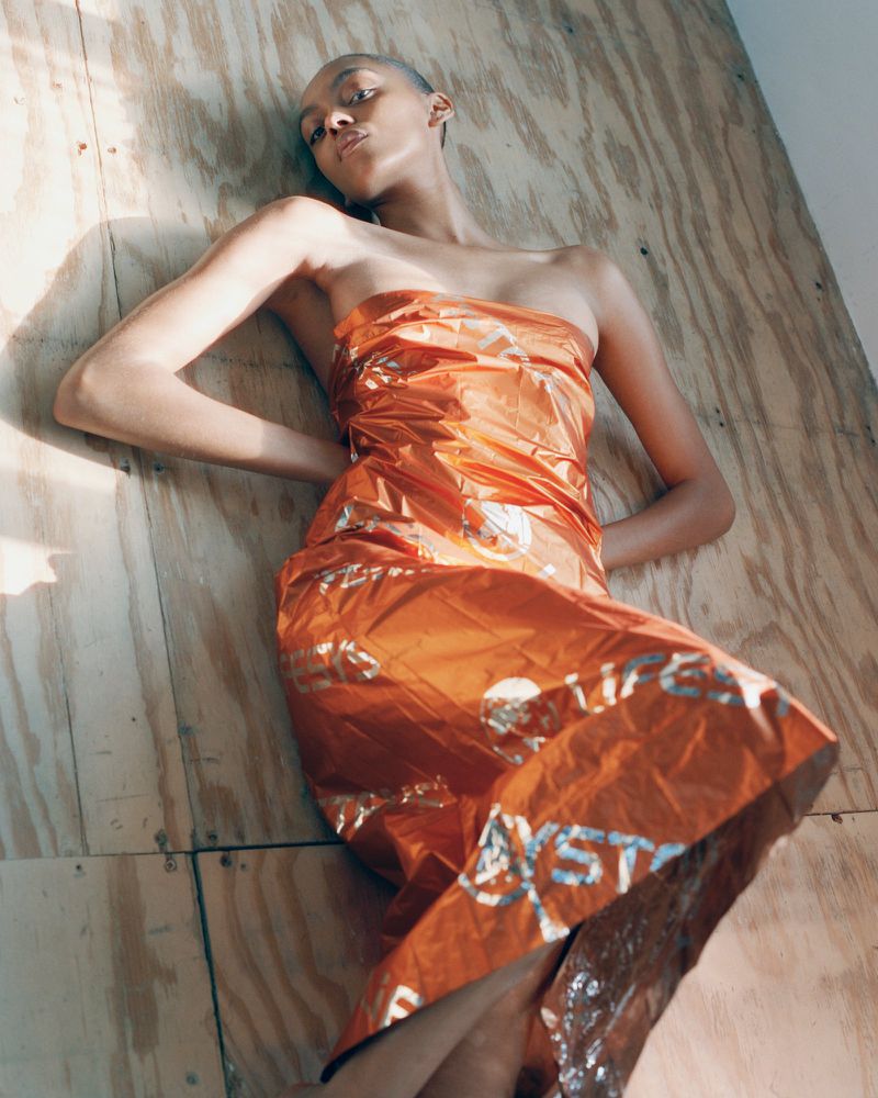 Model Bebe Parnell reclining in a strapless metallic orange dress with silver print on raw plywood boards under sunlight.