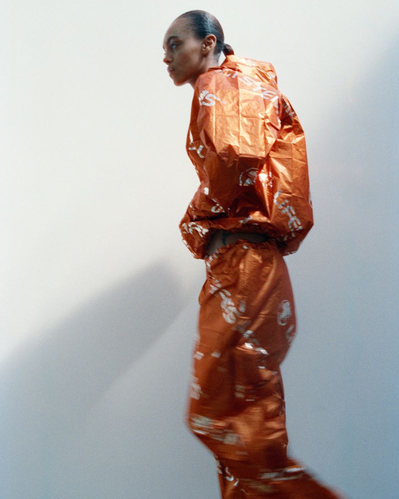 Model Bebe Parnell in a shiny metallic orange printed tracksuit against a plain studio background.