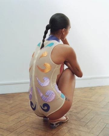 Model crouching on a herringbone wood floor wearing a sheer cream bodysuit with colorful swirl appliques and clear heels.