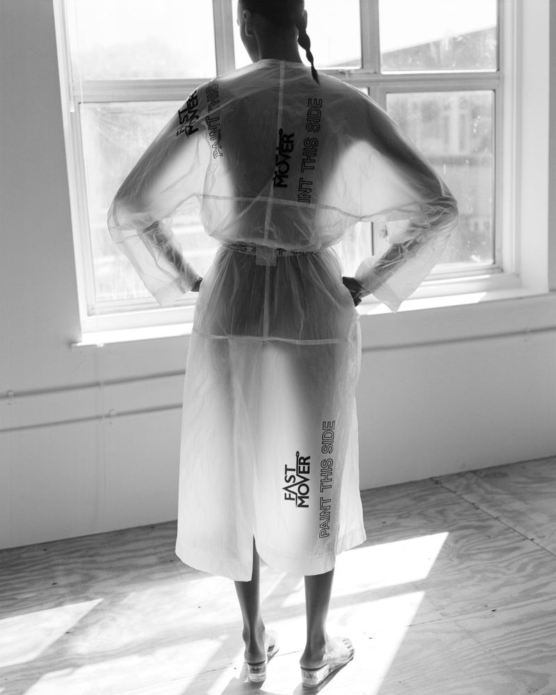 Model seen from behind wearing a translucent white printed coat and sandals standing by a large sunlit window in black and white.