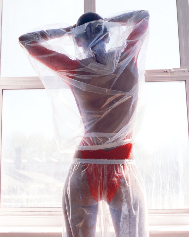 Model Bebe Parnell viewed from behind wearing a red bodysuit beneath a sheer plastic garment in front of a window.