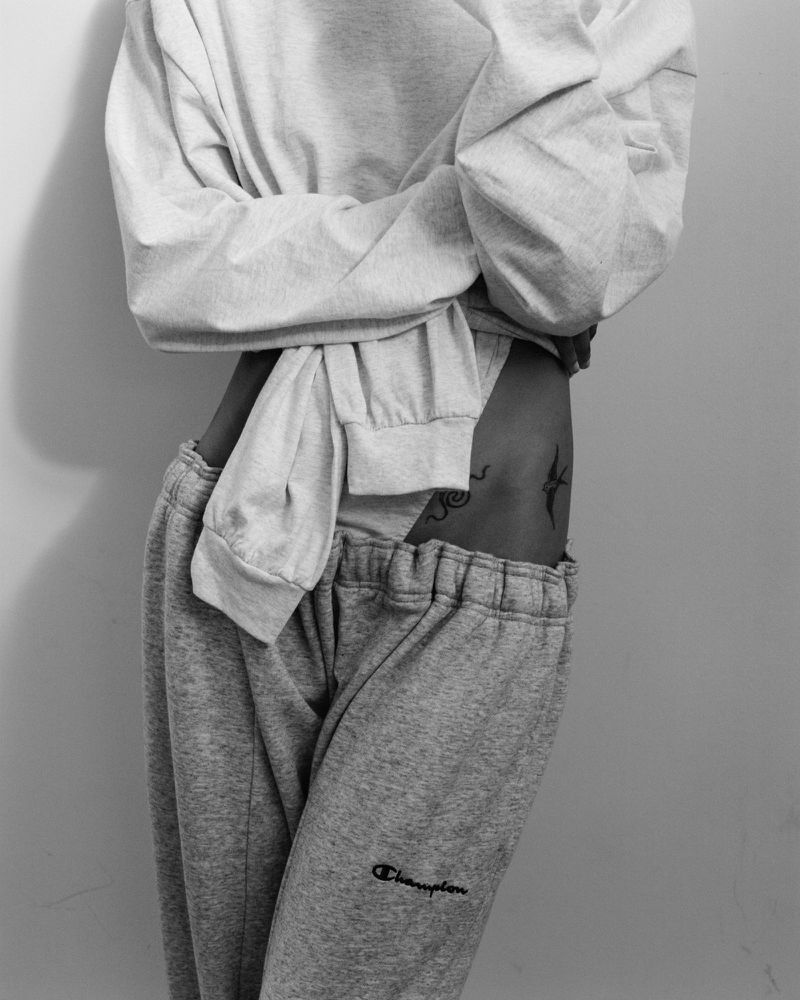 Monochrome crop of a model wearing a white long-sleeve top and low-slung grey sweatpants showing hip tattoos against a plain backdrop.