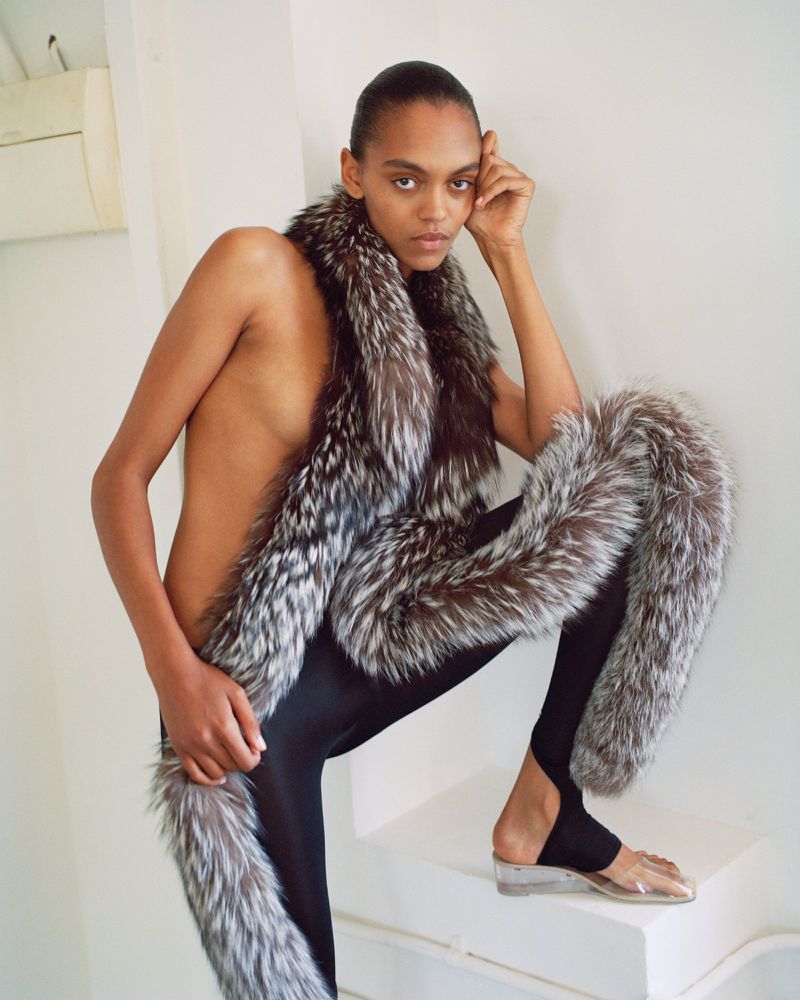 Model Bebe Parnell poses topless wrapped in a long fur boa with black stirrup leggings on white steps.