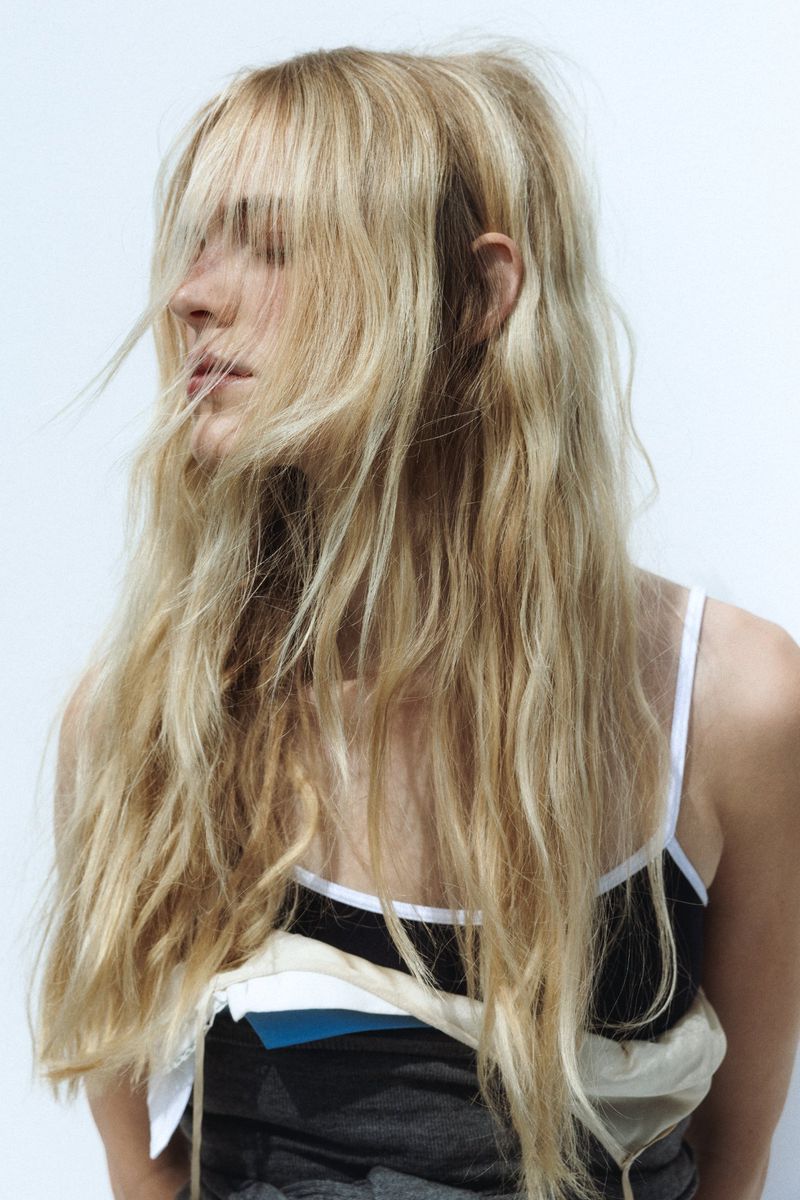 Close-up of a blonde woman with long wavy hair and a casual tank top against a white backdrop in a portrait style fashion photograph.