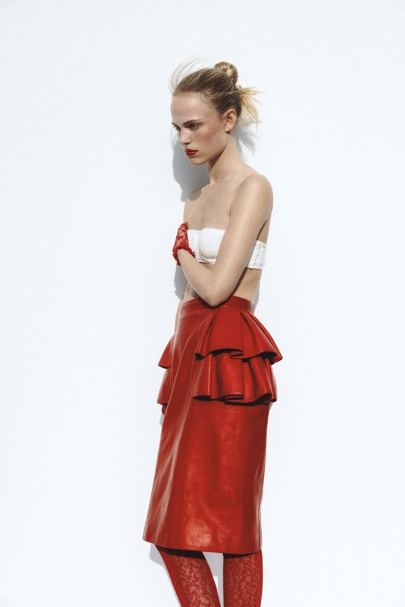 Blonde model in studio wears a white tube top, red leather ruffle skirt, and red patterned tights against a white background.