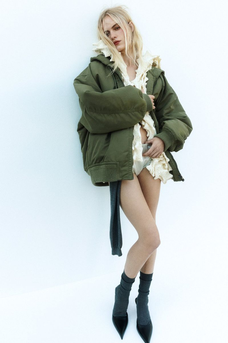 Fashion shot of a woman in an olive green jacket with a ruffled top and pumps posing in front of a white wall in a studio.