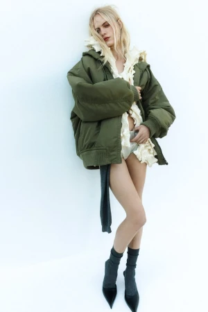 Fashion shot of a woman in an olive green jacket with a ruffled top and pumps posing in front of a white wall in a studio.